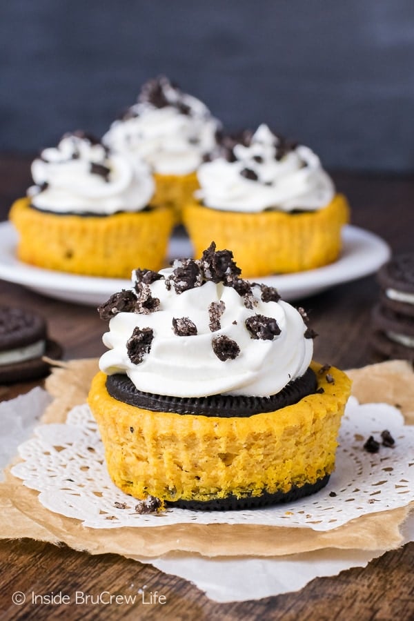 A pumpkin oreo cheesecake on a white doily with more cheesecakes behind it