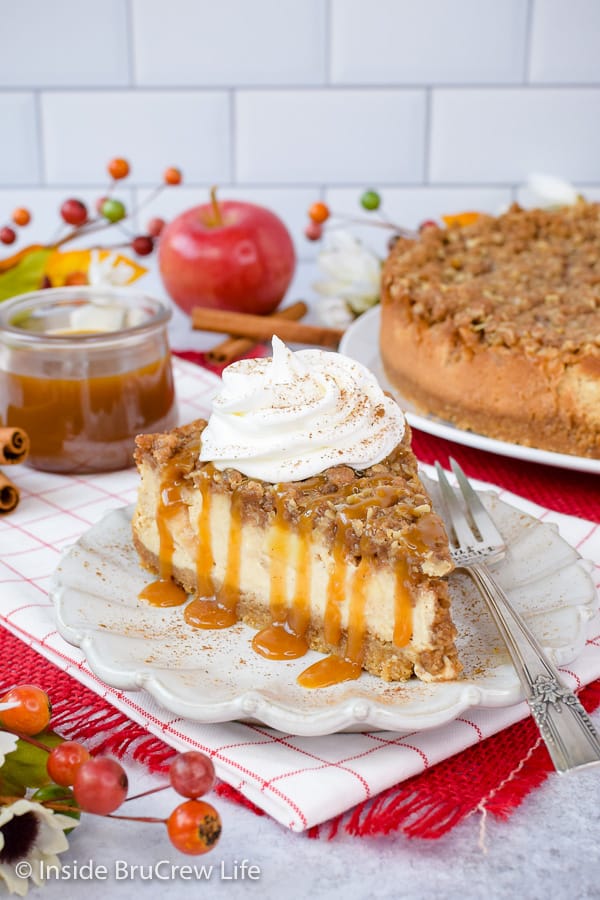 A slice of apple crisp cheesecake on a white plate with caramel drizzles and whipped cream on topl