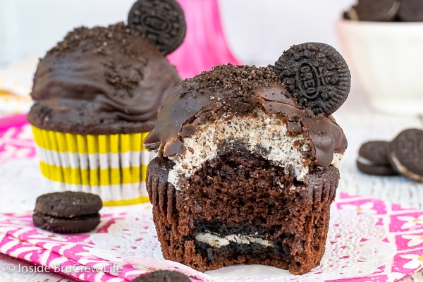 Two chocolate cupcakes topped with Oreo frosting, chocolate frosting, and a cookie with a bite missing out of the front one showing the hidden Oreo cookie