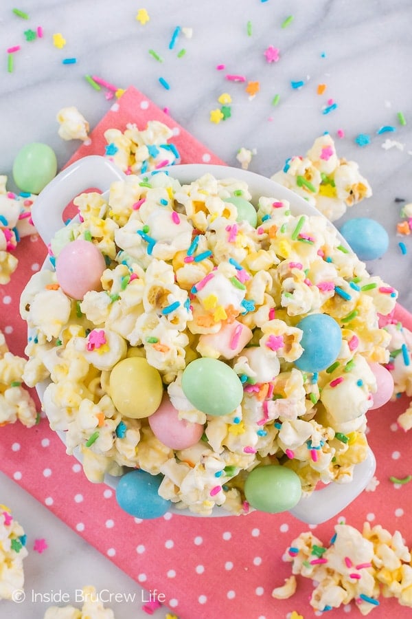 A white bowl on a pink towel filled with white chocolate popcorn loaded with sprinkles and candies.