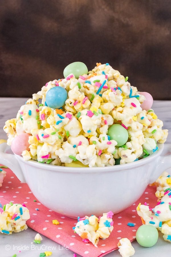 A white bowl on a pink towel filled with white chocolate popcorn that has sprinkles and candies in it.