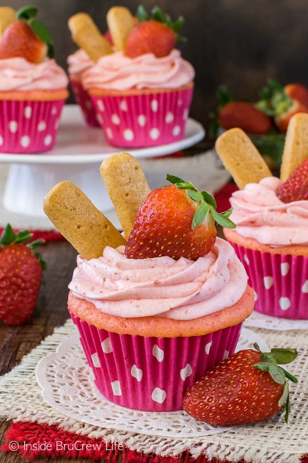 Two strawberry cheesecake cupcakes with strawberry frosting and a strawberry on doilies.