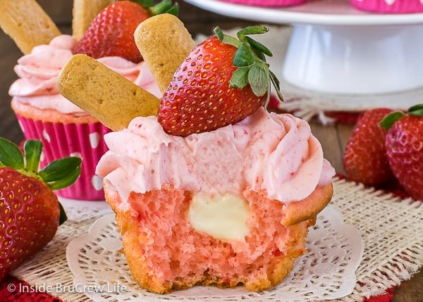 A white doily with a strawberry cupcake topped with strawberry frosting and a bite out of it showing the cheesecake center.