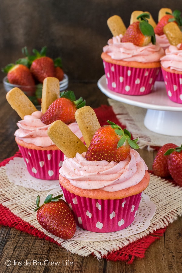 Two strawberry cheesecake cupcakes with strawberry frosting on doilies with a cake plate behind it.