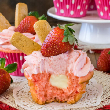 A strawberry cupcake on a white doily with a bite out of it showing the cheesecake center