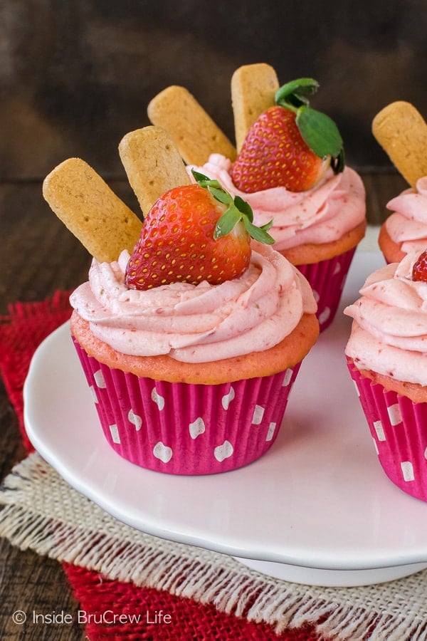 A white cake plate with strawberry cheesecake cupcakes on it.