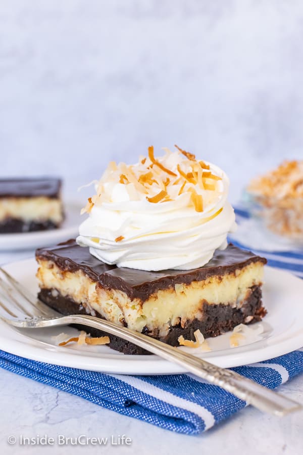 A square of Coconut Cream Cheesecake Brownies on a white plate with a swirl of Cool Whip and toasted coconut on top