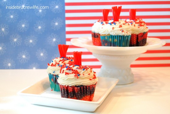 Firecracker cupcakes on a white plate and white cake stand with red white and blue papers behind them.