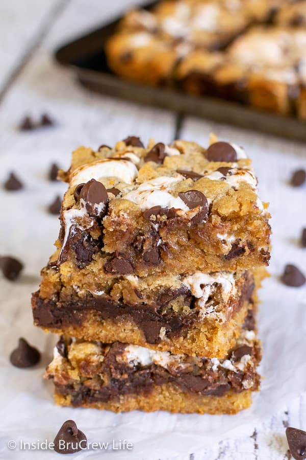 A stack of three Peanut Butter S'mores Blondies on a white background with a pan of blondies behind it
