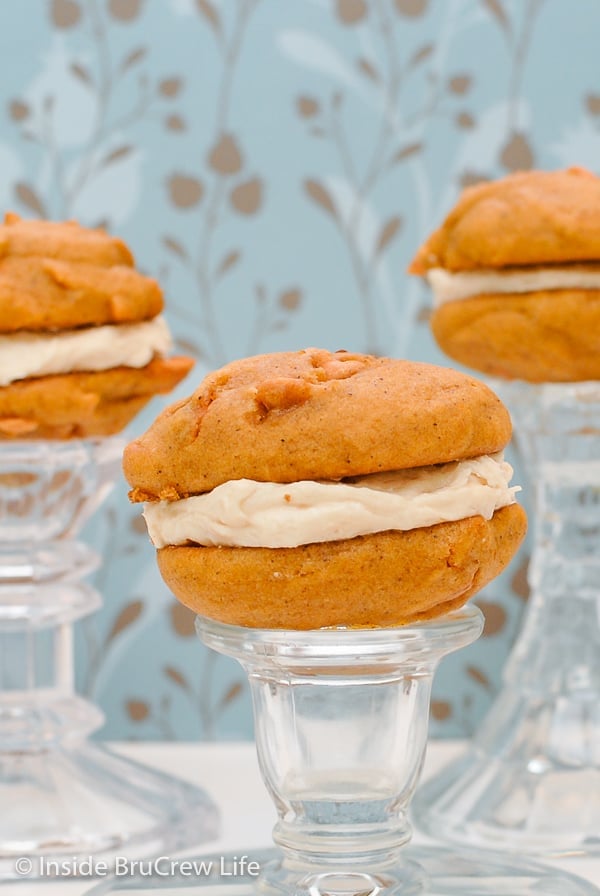 2 butterscotch pumpkin cookies with salted caramel frosting sandwiched in between them on a clear glass stand.