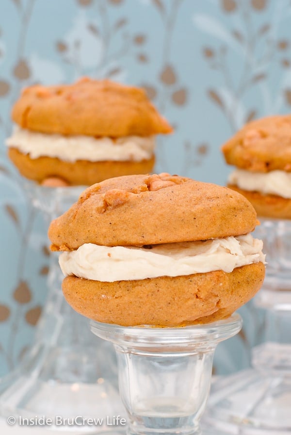 Close up of 2 butterscotch pumpkin cookies with salted caramel frosting sandwiched in between them on a clear glass stand.