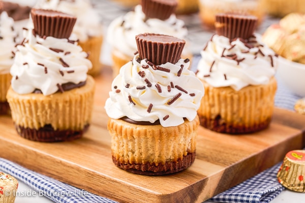 A wooden cutting board with mini peanut butter cheesecakes topped with chocolate, whipped cream, and peanut butter cups.