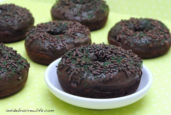 6 chocolate donuts with chocolate sprinkles and green sugar on a green background.