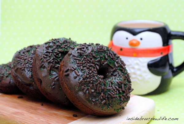 4 chocolate donuts on a wood cutting board with a penguin coffee mug in the background.