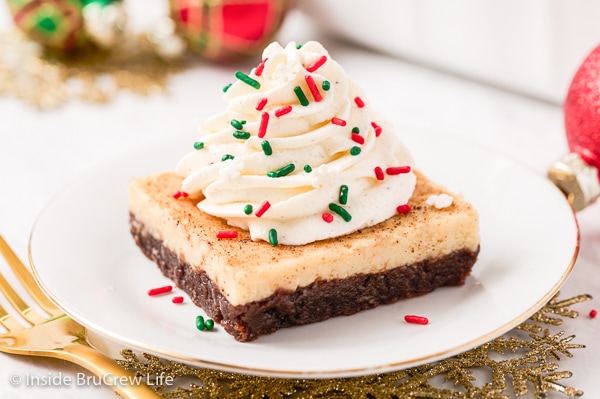 A white plate with a square of eggnog cheesecake brownies topped with whipped cream and sprinkles on it.