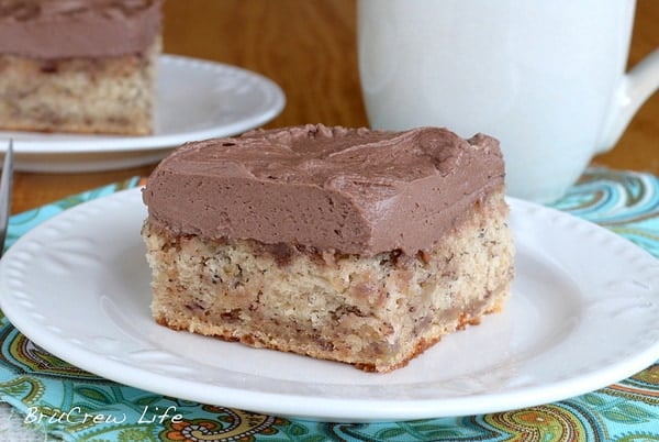 A white plate with a square of banana cake topped with chocolate frosting on it.