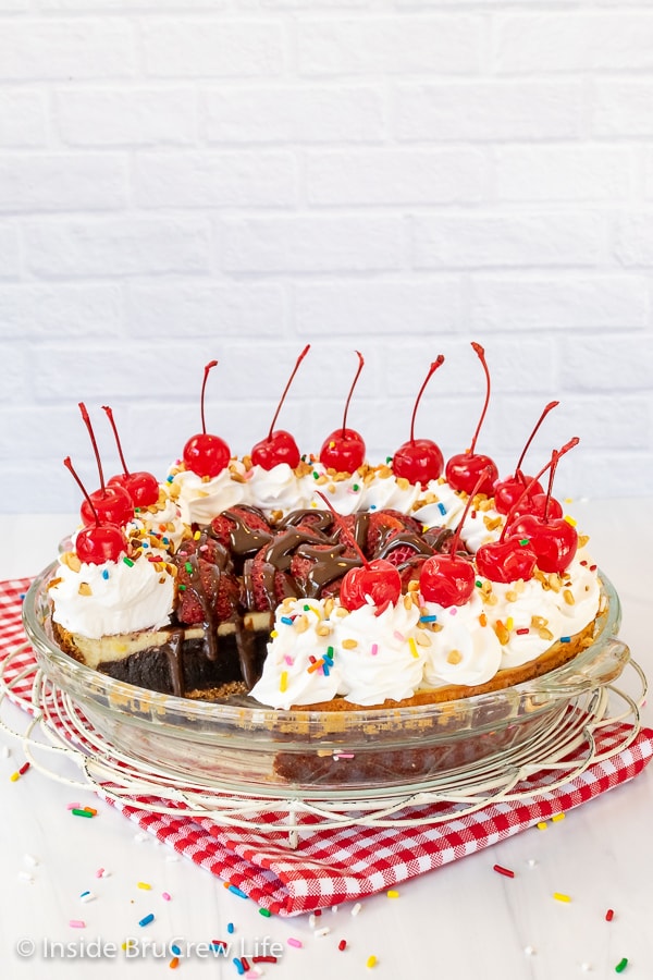 A white board with a glass pie plate filled with a brownie pie that is decorated with strawberries, cool whip, and cherries. 