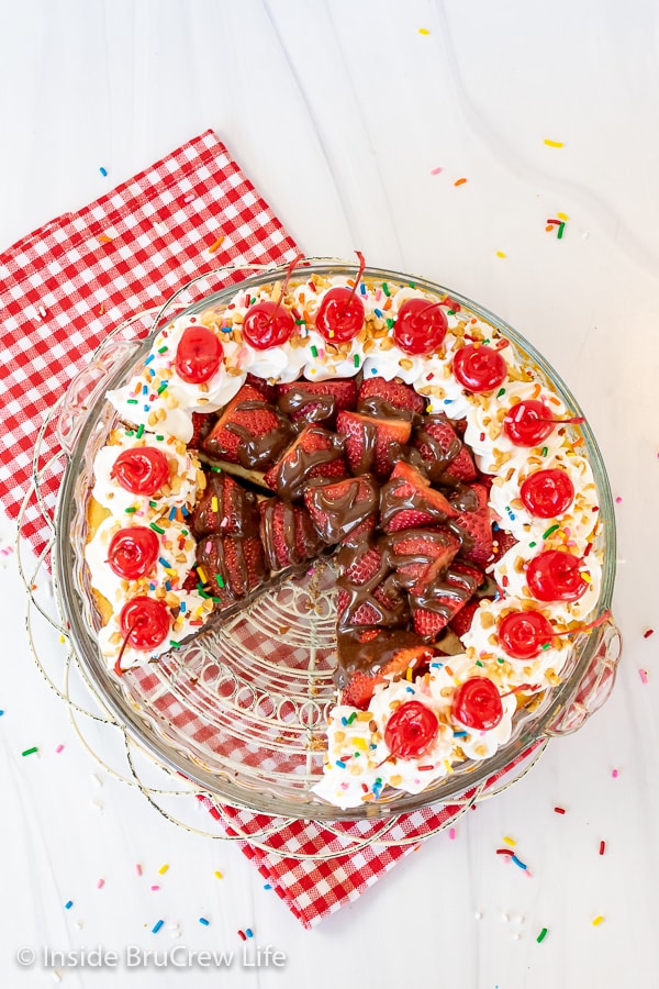 Overhead picture of a white board with a pie plate filled with a banana split pie topped with strawberries, cool whip, cherries, and sprinkles.