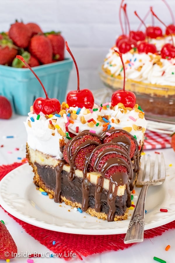 A white plate with a slice of banana split pie topped with strawberries, hot fudge, and toppings on it.