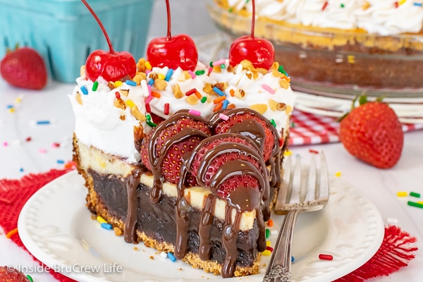 A white plate with a slice of brownie banana split pie with fruit and toppings on top.