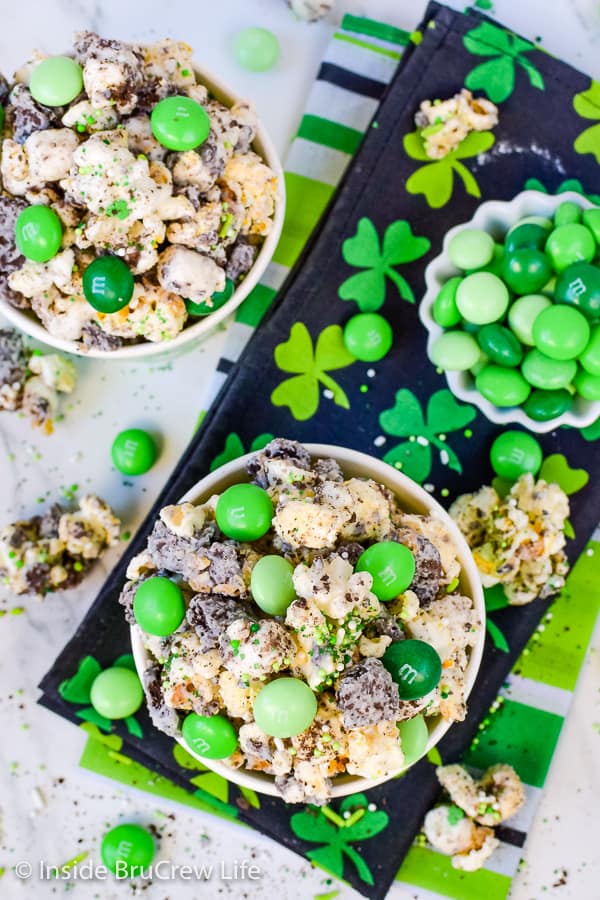 2 bowls filled with white chocolate popcorn mixed with green candies and mint cookies on a black towel with green clovers.