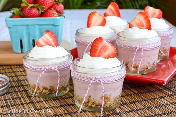 Six glass jars with pink and white ribbons filled with no bake strawberry cheesecake and topped with Cool Whip and strawberries