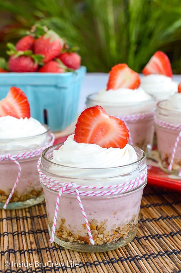 A small glass jar with a pink and white ribbon filled with no bake strawberry cheesecake and topped with Cool Whip and a fresh strawberry