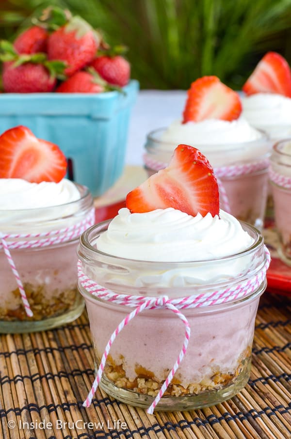 A clear glass jar filled with no bake strawberry cheesecake and topped with Cool Whip and a strawberry with more jars behind it