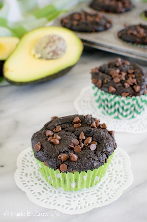 Two fudgy chocoolate avocado muffins on a white board with avocados and a muffin tin behind them