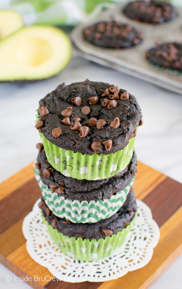 Three fudgy chocolate muffins with mini chocolate chips on top stacked on top of each other on a wooden board