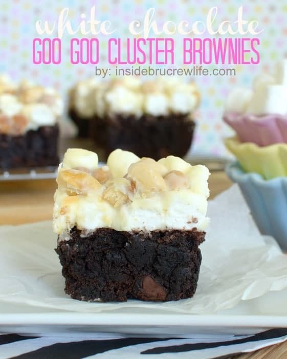 A white chocolate goo goo cluster brownie on a white plate with a few brownies in the background and a stack of pastel bowls with marshmallows to the side