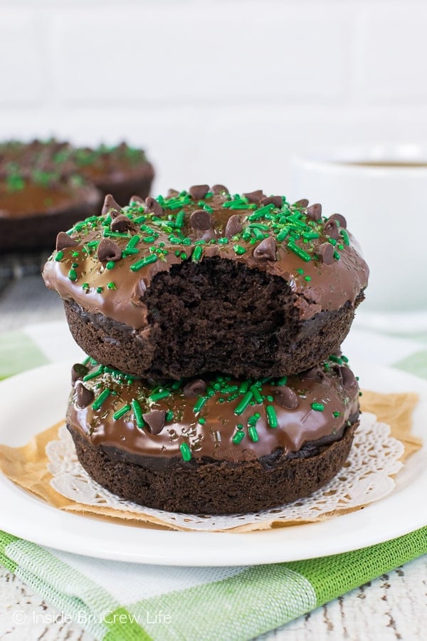 Two glazed baked chocolate donuts stacked on top of each other on a white plate with a bite out of the top one.