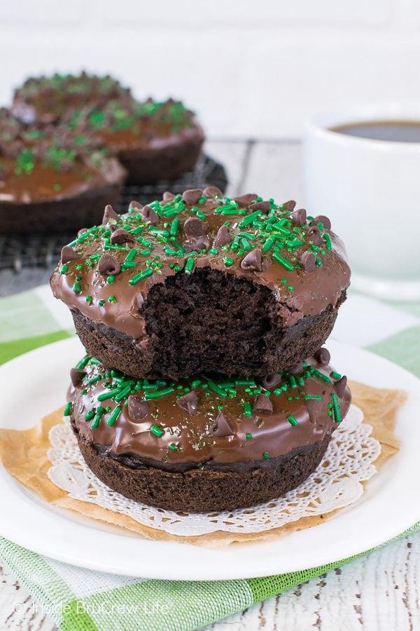 Two chocolate zucchini donuts topped with chocolate glaze and green sprinkles stacked on a white plate.