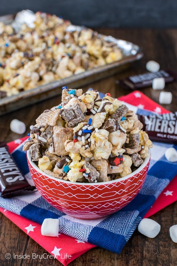 A red bowl filled with S'mores Popcorn with a tray behind it filled with more popcorn