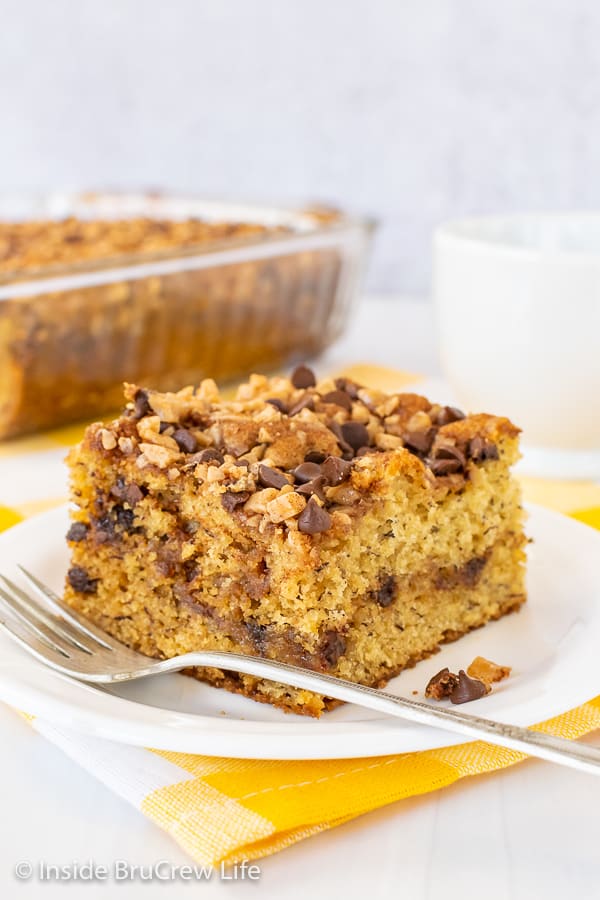 A slice of banana toffee cake on a white plate with a pan of banana cake behind it