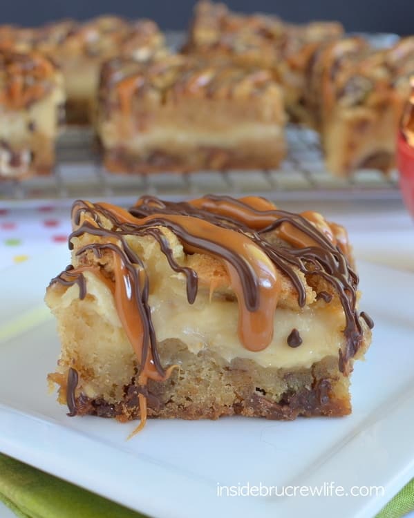 One Caramel Milky Way Cheesecake Bar on a white plate with a wire rack full of more cheesecake bars behind it