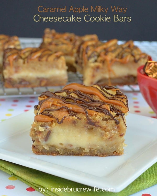 A square of Caramel Milky Way Cheesecake Cookie Bars on a white plate with a wire rack behind it with more cookie bars on it