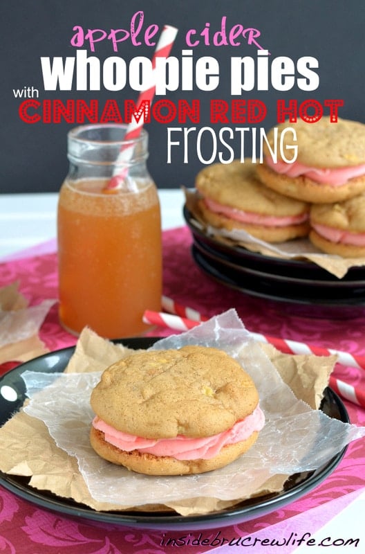 A black plate with a apple cider whoopie pie filled with cinnamon red hot frosting on it and a glass of apple cider behind it