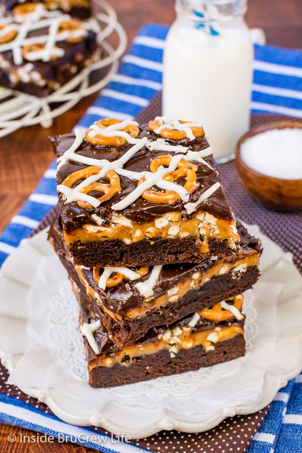 Three salted caramel brownies stacked on a plate with white chocolate drizzles and pretzels on top.