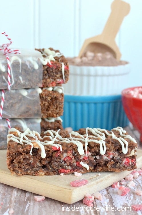 A peppermint hot chocolate granola bar on a wood tray with white chocolate drizzles and bowls of ingredients behind it