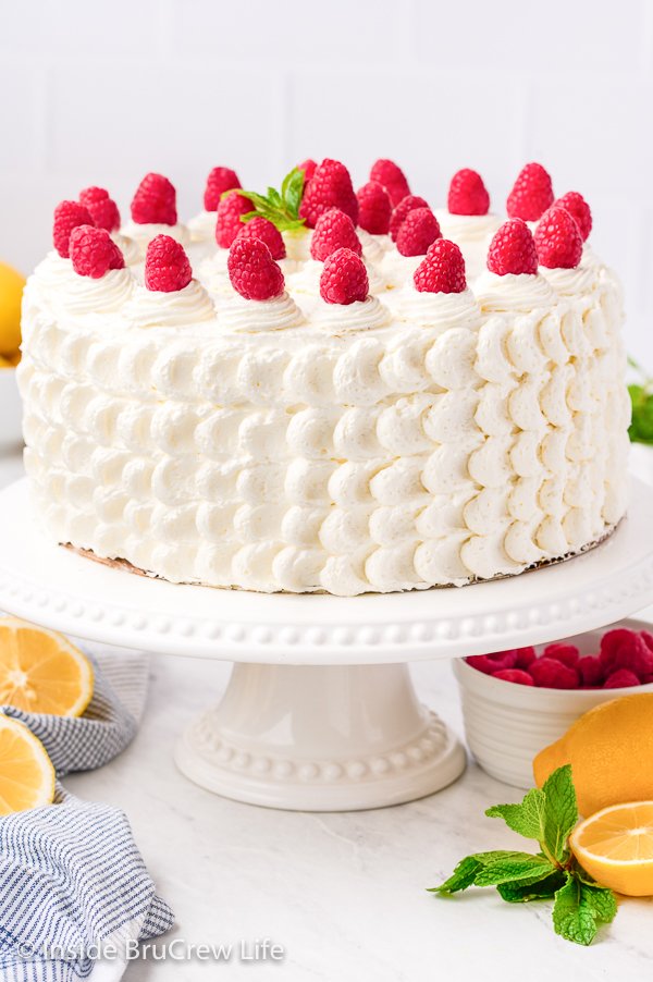 A white cake plate with a frosted cake topped with raspberries on it.