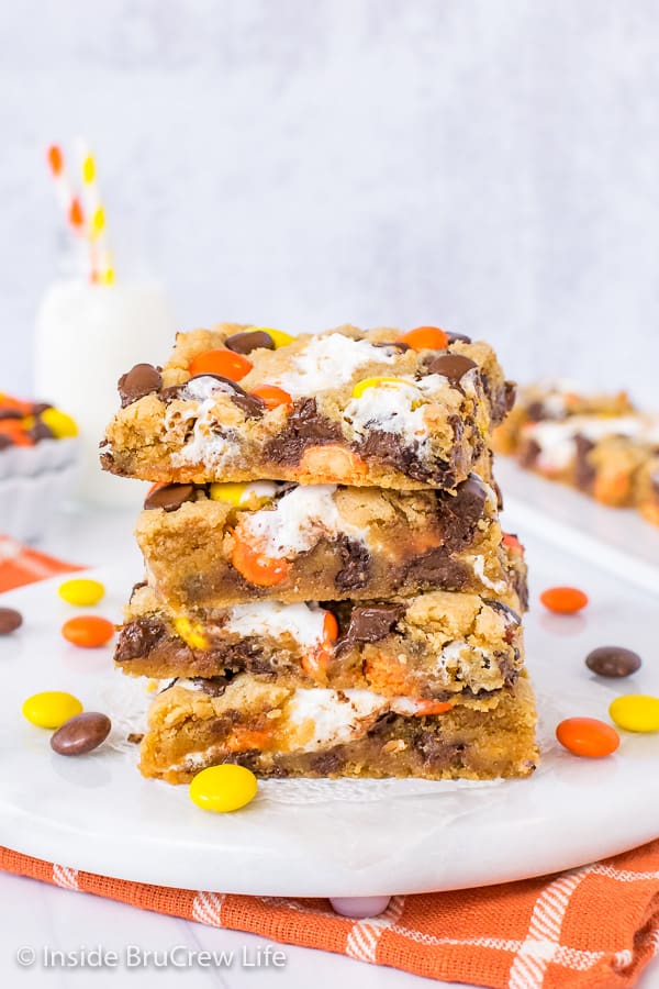 Four squares of peanut butter blondies stacked up on a white board with Reese's Pieces scattered around it