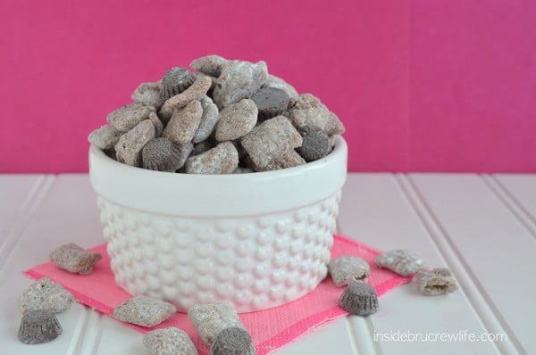 A white bowl of powdered sugar coated chex mix with chocolates.