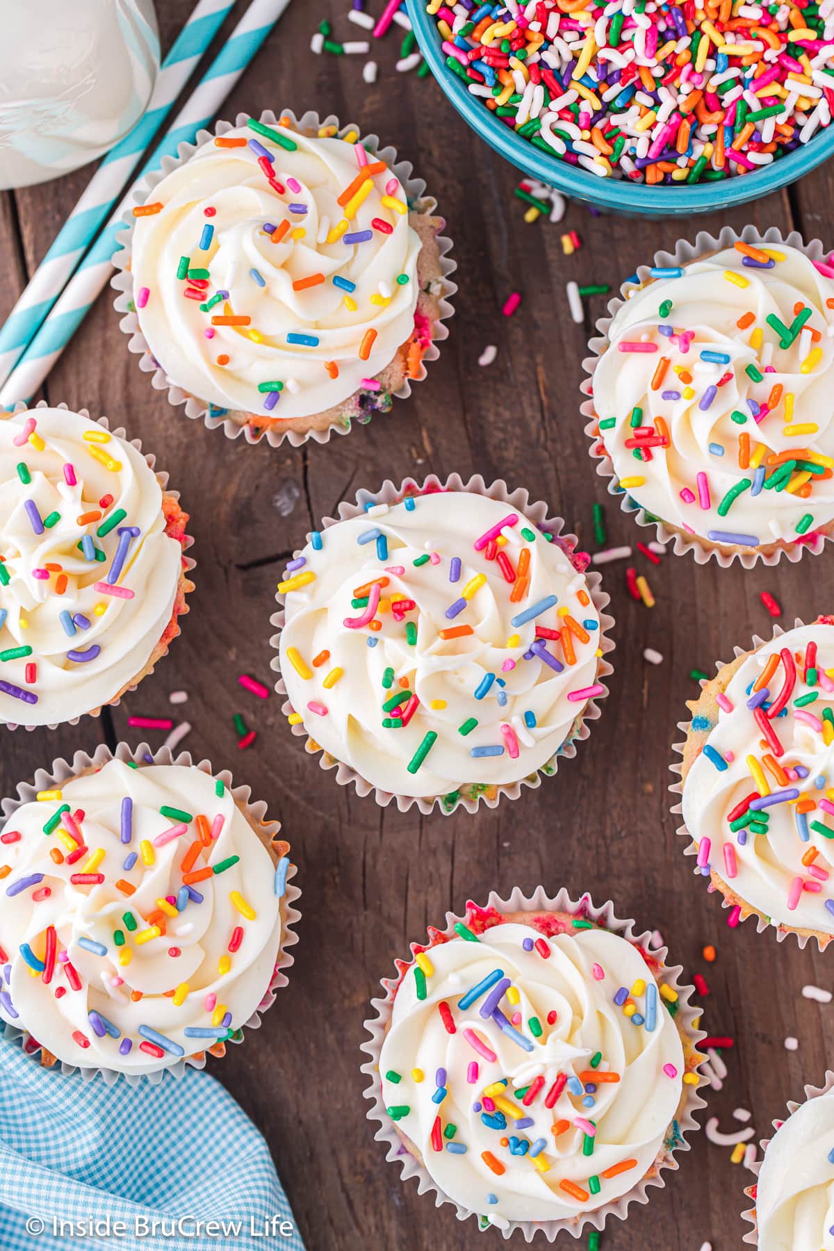 Frosted birthday cake cupcakes topped with sprinkles on a brown board.