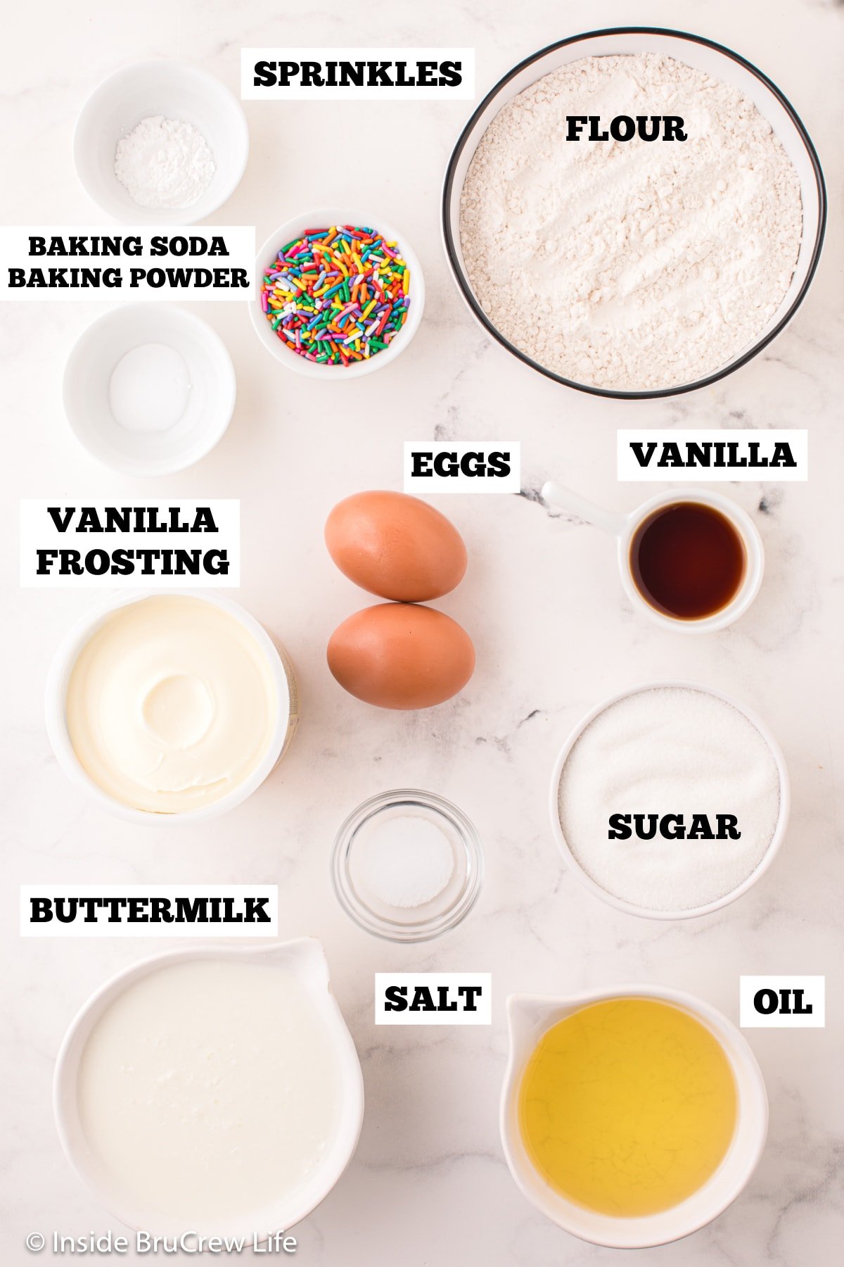 A white board with bowls of ingredients needed to make vanilla cupcakes with sprinkles.