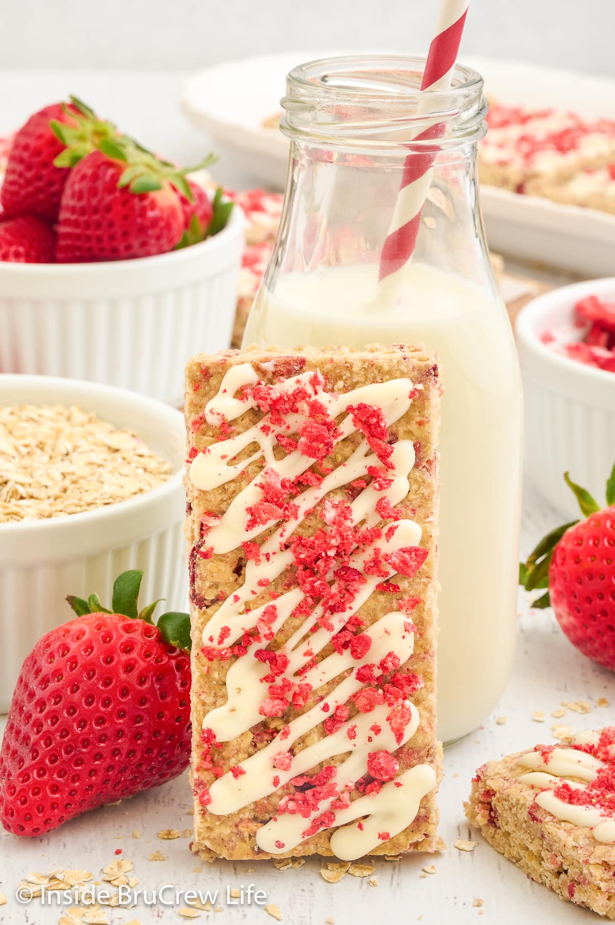A strawberry topped granola bar standing next to a glass of milk.