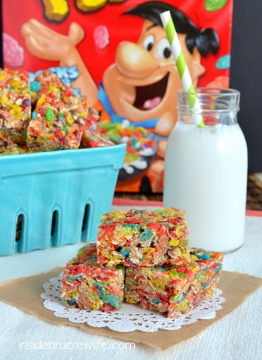 A stack of fruity pebbles granola bars stacked together on a white doily with a box of cereal behind them