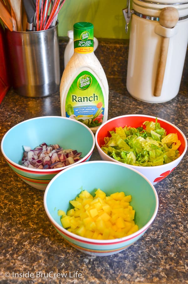 Three bowls filled with chopped mangos, onions, and lettuce and a bottle of ranch dressing behind them