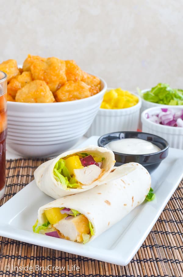 A white plate with a mango chicken wrap cut in half on it and bowls of toppings behind it