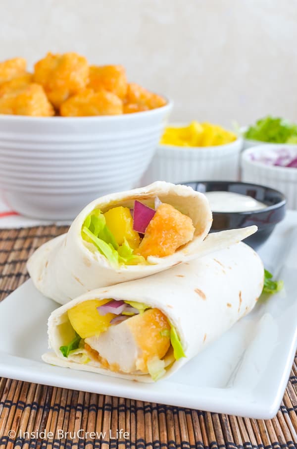 A white plate with a mango chicken wrap cut in half and leaning on its side and bowls of toppings behind it